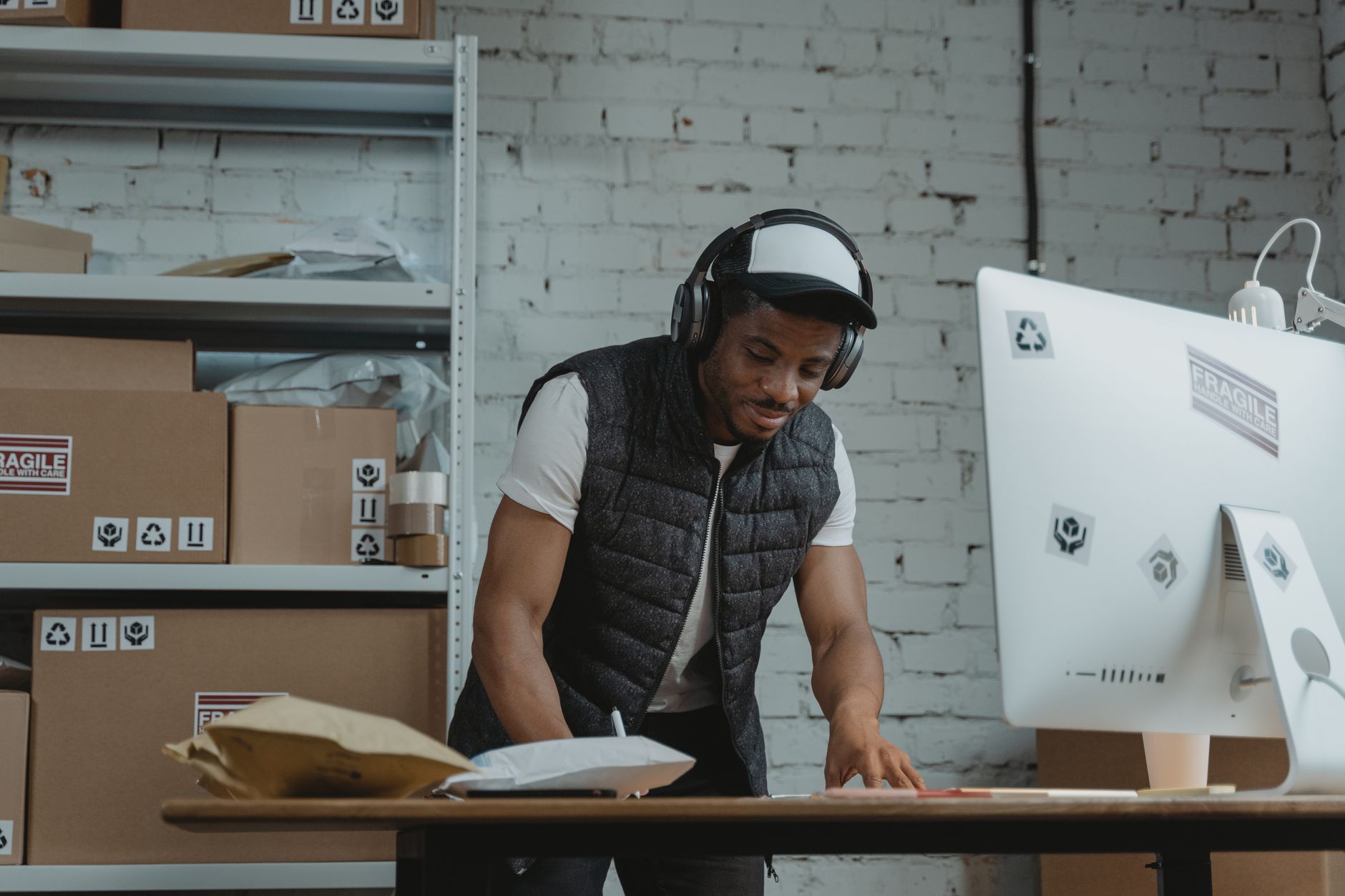 Businessman in a warehouse standing by computer, wearing headphones and vest, managing packages.